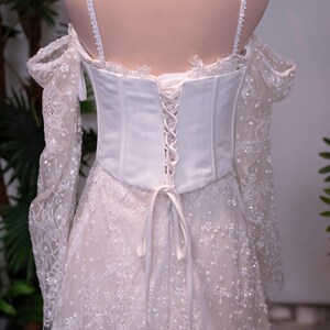 May include: A white off-the-shoulder gown featuring a corset-style bodice with lace-up back closure, thin spaghetti straps, and long, sheer lace sleeves. The dress is adorned with intricate floral lace patterns and small, scattered white beads.