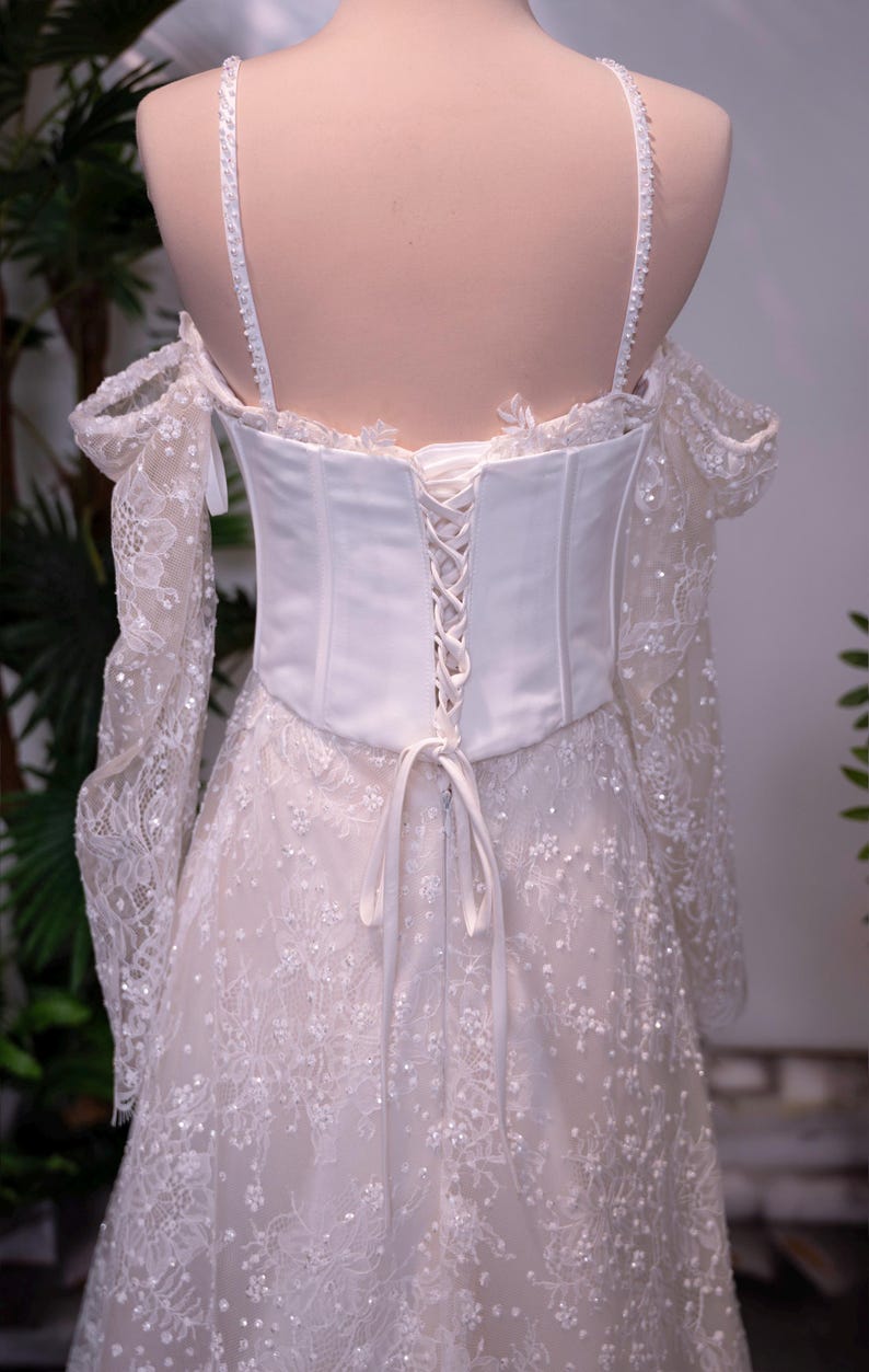 May include: A white off-the-shoulder gown featuring a corset-style bodice with lace-up back closure, thin spaghetti straps, and long, sheer lace sleeves. The dress is adorned with intricate floral lace patterns and small, scattered white beads.