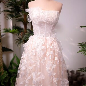 May include: A strapless, blush-colored wedding dress on a mannequin. The bodice is embellished with pearls and a floral applique. The skirt features delicate, white floral embroidery. A gold crown sits atop the mannequin's head.