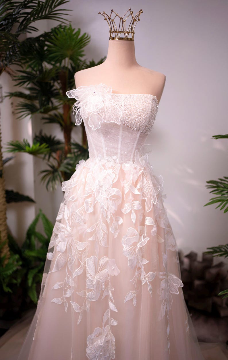 May include: A strapless, blush-colored wedding dress on a mannequin. The bodice is embellished with pearls and a floral applique. The skirt features delicate, white floral embroidery. A gold crown sits atop the mannequin's head.