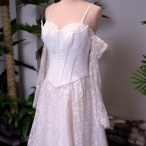 May include: A white corset-style dress featuring a structured bodice with thin straps, off-the-shoulder lace sleeves, and a floor-length skirt covered in intricate floral lace embroidery with subtle sequin detailing, topped with a gold crown.