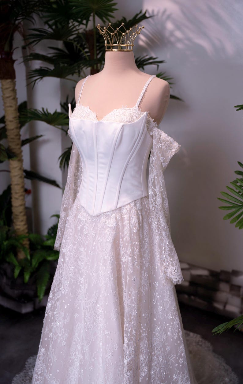 May include: A white corset-style dress featuring a structured bodice with thin straps, off-the-shoulder lace sleeves, and a floor-length skirt covered in intricate floral lace embroidery with subtle sequin detailing, topped with a gold crown.