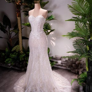 May include: A strapless, white wedding dress with a mermaid silhouette. The gown is embellished with sequins and lace, and it has a long train. A gold crown sits atop the mannequin's head. The dress is displayed in front of tropical plants.