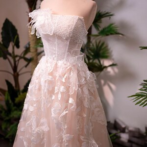 May include: A strapless, blush-colored wedding gown on a mannequin. The bodice is embellished with pearls and the skirt features floral appliqu&eacute;s. A gold crown sits atop the mannequin's head. The dress is displayed in front of greenery.
