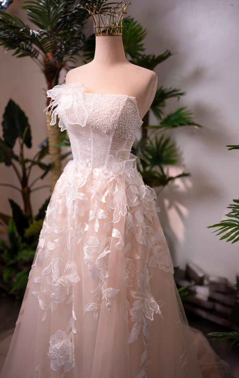 May include: A strapless, blush-colored wedding gown on a mannequin. The bodice is embellished with pearls and the skirt features floral appliqu&eacute;s. A gold crown sits atop the mannequin's head. The dress is displayed in front of greenery.