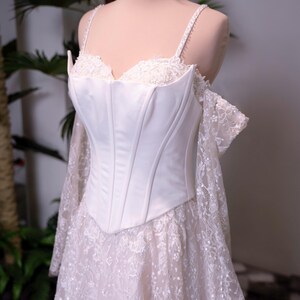 May include: A white corset-style bodice dress featuring a sweetheart neckline, thin spaghetti straps, and off-the-shoulder lace sleeves. The skirt is made of intricate white floral lace with subtle sequin detailing, and the bodice is smooth satin.