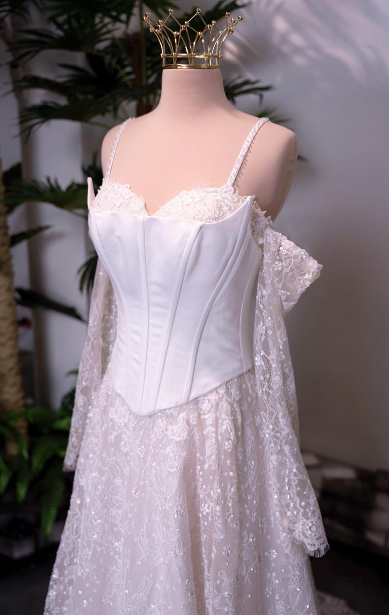 May include: A white corset-style bodice dress featuring a sweetheart neckline, thin spaghetti straps, and off-the-shoulder lace sleeves. The skirt is made of intricate white floral lace with subtle sequin detailing, and the bodice is smooth satin.