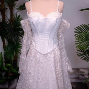 May include: A white corset-style bodice dress featuring thin straps, a sweetheart neckline, and off-the-shoulder long lace sleeves. The skirt is made of intricate white floral lace with subtle sparkle, and a gold-toned crown sits atop the mannequin.