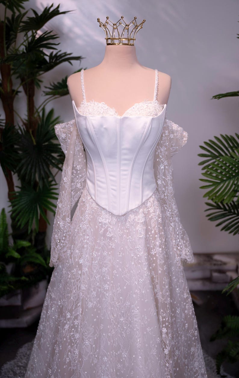 May include: A white corset-style bodice dress featuring thin straps, a sweetheart neckline, and off-the-shoulder long lace sleeves. The skirt is made of intricate white floral lace with subtle sparkle, and a gold-toned crown sits atop the mannequin.