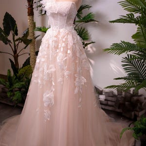 May include: A strapless blush-colored wedding gown on a mannequin. The bodice is embellished with beads and lace, and the skirt features delicate floral appliqu&eacute;s. A gold crown sits atop the mannequin's head. The dress has a flowing, layered design.