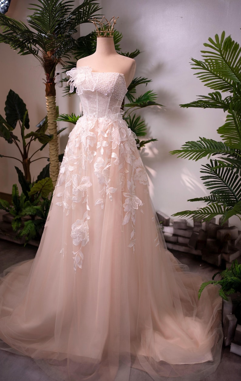 May include: A strapless blush-colored wedding gown on a mannequin. The bodice is embellished with beads and lace, and the skirt features delicate floral appliqu&eacute;s. A gold crown sits atop the mannequin's head. The dress has a flowing, layered design.