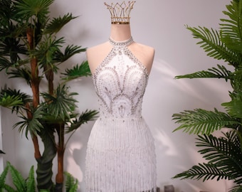 Könnte beinhalten: Ein weißes Minikleid mit hohem Neckholder-Ausschnitt und aufwendigen silbernen Kristallverzierungen, die in einem dekorativen Muster über das Oberteil angeordnet sind. Der Rock ist mit langen, herabhängenden weißen Perlenfransen besetzt.