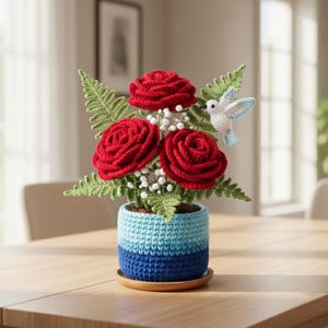 May include: A handcrafted crochet floral arrangement featuring three red roses, green fern leaves, and a small white hummingbird with blue wings. The flowers are in a blue and light blue striped pot, sitting on a wooden saucer.