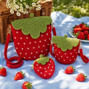 Puede incluir: Un conjunto de tres bolsos y un monedero de ganchillo con temática de fresa. Los bolsos son rojos con detalles de semillas blancas y copas de hojas verdes. Una cesta de picnic y fresas frescas en el fondo.