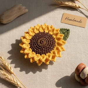 Puede incluir: Un girasol hecho a mano con ganchillo con pétalos amarillos, un centro marrón y una hoja verde. El centro tiene pequeñas cuentas doradas. Una pequeña etiqueta marrón con la palabra "Handmade" es visible.