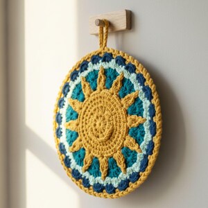 May include: A round, handmade crochet potholder with a sunburst design. The center is mustard yellow, surrounded by teal, light blue, and navy blue rings. A loop is attached for hanging. The potholder is hanging on a wooden peg.