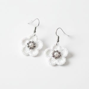 Pode incluir: Um par de brincos de flores brancas de crochê. Cada brinco apresenta um design de flor de cinco pétalas com um centro de pérolas e contas prateadas. Os brincos penduram-se em ganchos de orelha prateados.