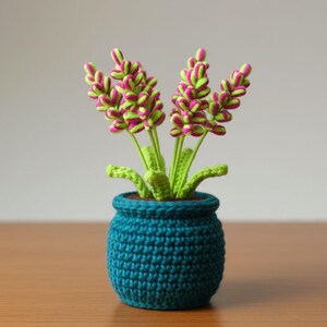May include: A handmade crochet plant in a teal pot. The plant features green stems and leaves, with vibrant pink and green flower heads. The pot is round and textured, sitting on a wooden surface.