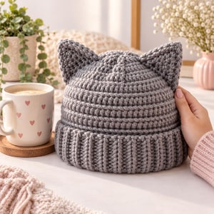 Puede incluir: Un gorro de ganchillo gris que presenta dos orejas de gato puntiagudas en la parte superior. El gorro está construido con un borde acanalado y un patrón de puntos texturizados en todo el cuerpo principal. El diseño es simple y funcional, mostrando la textura.