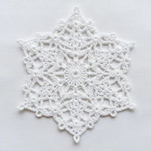 Może przedstawiać: Biała, szydełkowa serwetka w kształcie płatka śniegu. Wzór płatka śniegu charakteryzuje się centralnym okrągłym wzorem z promienistymi elementami kwiatowymi i geometrycznymi. Krawędzie są karbowane, tworząc delikatny, koronkowy wygląd. Serwetka wykonana jest z białej przędzy.