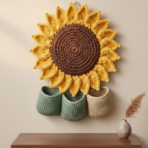 Può includere: Un grande arazzo da parete a forma di girasole fatto a mano all'uncinetto, con petali gialli e un centro marrone. Sotto il girasole ci sono tre piccoli cestini all'uncinetto attaccati in tonalità di verde e beige. Il pezzo è montato su una parete chiara.