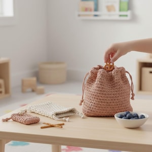 May include: A collection of handmade crochet bags in various sizes and colors. The main bag is a dusty rose color, with a drawstring closure. A hand is placing a pretzel into the bag. Other bags are off-white and dusty rose. A small bowl of blueberries is also on the table.