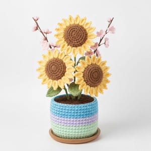 May include: A handmade crochet sunflower arrangement in a striped blue, lilac, and green pot. The sunflowers have yellow petals and brown centres, with green leaves and pink flower accents. The pot sits on a wooden saucer.