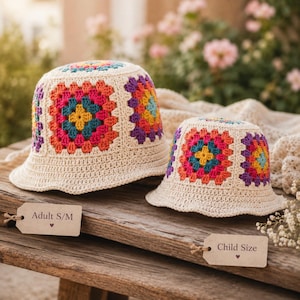 May include: Two crochet bucket hats, one adult size and one child size, sit on a wooden surface. The hats are cream-colored with colorful square patterns in the center. The adult hat has a tag that reads "Adult S/M" and the child hat has a tag that reads "Child Size."
