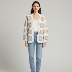 Puede incluir: Un cárdigan de crochet crema con un patrón floral en tonos pastel. El cárdigan abierto presenta un borde festoneado y se combina con una camiseta blanca y unos vaqueros azules. El estilo es bohemio y hecho a mano.