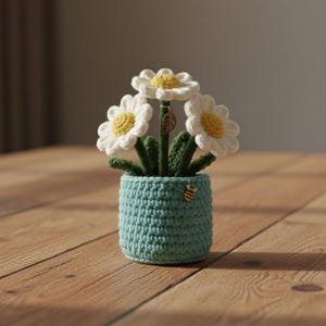 Pode incluir: Um arranjo de flores de margarida de crochê em um vaso azul-turquesa. As margaridas têm pétalas brancas e centros amarelos. O vaso tem um pequeno pingente de abelha preso. O arranjo está em uma superfície de madeira.