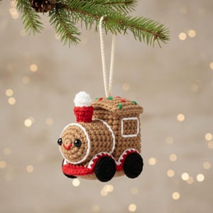 Puede incluir: Adorno navideño tejido a ganchillo con forma de tren marrón con detalles rojos y blancos. El tren tiene ruedas negras, una chimenea roja y una cuerda blanca para colgarlo. El adorno está suspendido de una rama de pino verde.