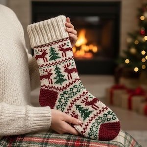 Peut inclure: Une chaussette de Noël tricotée avec un revers et un corps de couleur crème. La chaussette présente un motif de rennes rouges et de sapins verts sur un fond crème. Le bout et le talon sont de couleur rouge foncé.