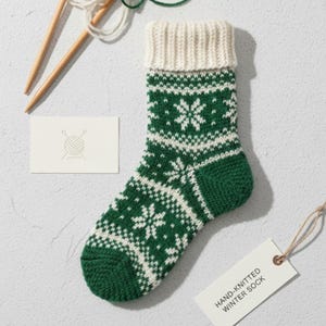 Könnte beinhalten: Handgestrickte Wintersocke mit weißen Schneeflocken auf grünem Grund. Der Bund ist cremefarben. Stricknadeln, Garn und eine Karte mit Wollknäuel-Logo sind im Bild. Ein Etikett trägt die Aufschrift "HAND-KNITTED WINTER SOCK."