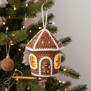 May include: A crocheted gingerbread house Christmas ornament with a white roof, brown walls, and colorful sprinkles. The ornament has a white string for hanging and is illuminated from within. It hangs on a decorated Christmas tree.