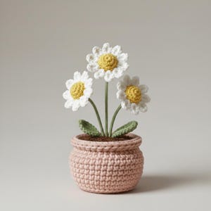 Puede incluir: Un arreglo de flores de margaritas de ganchillo en una maceta rosa. Las margaritas tienen pétalos blancos y centros amarillos. La maceta es de color rosa claro y tiene un aspecto texturizado. El arreglo incluye hojas y tallos verdes.