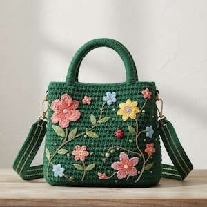 Könnte beinhalten: Grüne Häkeltasche mit floralem Design. Die Tasche hat einen oberen Griff und einen abnehmbaren Schultergurt. Die Blumen sind in Rosa-, Gelb- und Blautönen gehalten, mit goldenen Akzenten und einem kleinen Marienkäfer. Die Tasche steht auf einer Holzoberfläche.