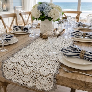 May include: A cream-colored crochet table runner with a scalloped edge, set on a wooden dining table with white plates, blue and white striped napkins in woven rings, clear crystal glassware, and a white vase filled with blue and white hydrangeas.