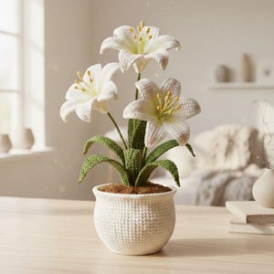 May include: A crocheted white lily plant in a white pot. The plant features three large, open lilies with yellow stamens and green leaves. The pot has a textured, woven appearance and is filled with brown material.