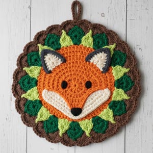 May include: A round, crocheted fox-faced hot pad. The fox has orange fur, black eyes, and a white muzzle. The pad is surrounded by green leaves and a brown scalloped border. A loop is attached for hanging.
