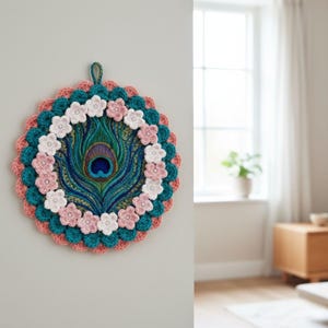 May include: A handcrafted peacock feather wall hanging. The circular design features a central peacock feather, surrounded by teal, coral, and white crochet details. Pink and white crochet flowers are arranged around the feather, adding a decorative touch.