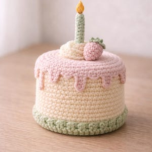 Puede incluir: Una tarta de cumpleaños de ganchillo con una vela y una fresa. La tarta tiene una base amarilla clara, glaseado rosa con goteo y una base verde. La vela es verde con una llama amarilla. Una fresa rosa está encima.