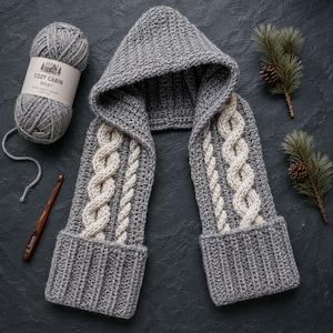 May include: A gray hooded scarf featuring a textured knit pattern with cream-colored cable knit accents on the front panels. The scarf includes a hood and wide ribbed cuffs at the ends. A ball of gray yarn labeled Cozy Cabin Bulky and a wooden crochet hook.