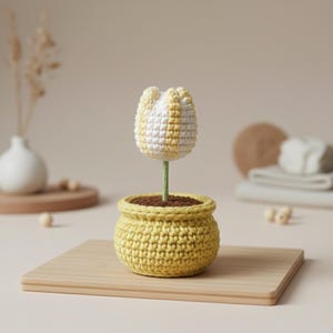 Könnte beinhalten: Eine handgefertigte Häkeltulpe in einem gelben Topf. Die Tulpenblume ist weiß und gelb, mit einem grünen Stiel. Der Topf ist gelb und mit brauner Erde gefüllt. Die Häkelblume steht auf einem Holzbrett.