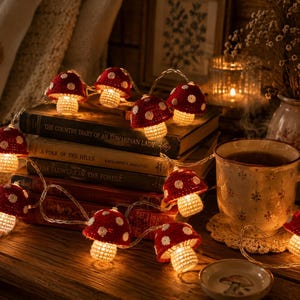 Pode incluir: Um fio de luzes LED com cogumelos de croché com chapéus vermelhos de pintas brancas e caules cor de creme. As luzes estão dispostas sobre uma pilha de livros antigos, com cada cogumelo a brilhar por dentro, emitindo uma luz quente.