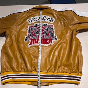 May include: A mustard yellow jacket with white shoulder panels and a ribbed hem featuring yellow, white, and dark brown stripes. The back displays a graphic with two dragons and the text "DRAGONS" and "AVIREX" next to a measuring tape showing 30 inches.