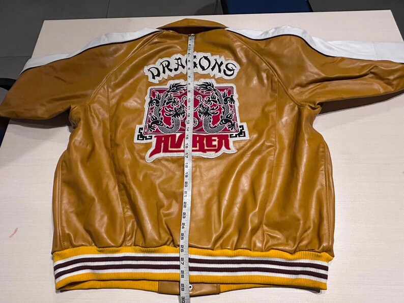 May include: A mustard yellow jacket with white shoulder panels and a ribbed hem featuring yellow, white, and dark brown stripes. The back displays a graphic with two dragons and the text "DRAGONS" and "AVIREX" next to a measuring tape showing 30 inches.