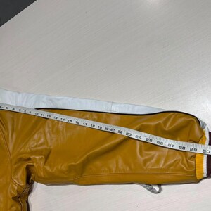 May include: A mustard yellow varsity jacket sleeve with a white leather stripe along the top. The sleeve features a ribbed yellow, white, and maroon cuff. A measuring tape is placed along the sleeve, showing a length of approximately 32 inches.