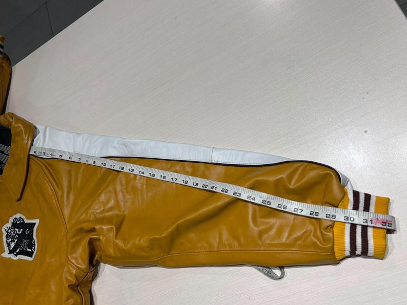 May include: A mustard yellow varsity jacket sleeve with a white leather stripe along the top. The sleeve features a ribbed yellow, white, and maroon cuff. A measuring tape is placed along the sleeve, showing a length of approximately 32 inches.