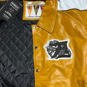 May include: A mustard yellow and white varsity jacket featuring a black quilted interior lining, silver snap buttons, and a black embroidered letter A patch. The inner label reads AVIREX AUTHENTIC VARSITY SPORTS EQUIPMENT AVIREX U.S.A. NEW YORK.