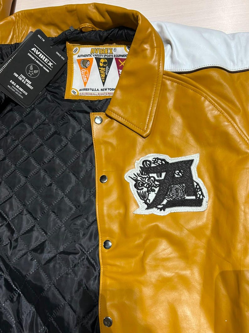 May include: A mustard yellow and white varsity jacket featuring a black quilted interior lining, silver snap buttons, and a black embroidered letter A patch. The inner label reads AVIREX AUTHENTIC VARSITY SPORTS EQUIPMENT AVIREX U.S.A. NEW YORK.